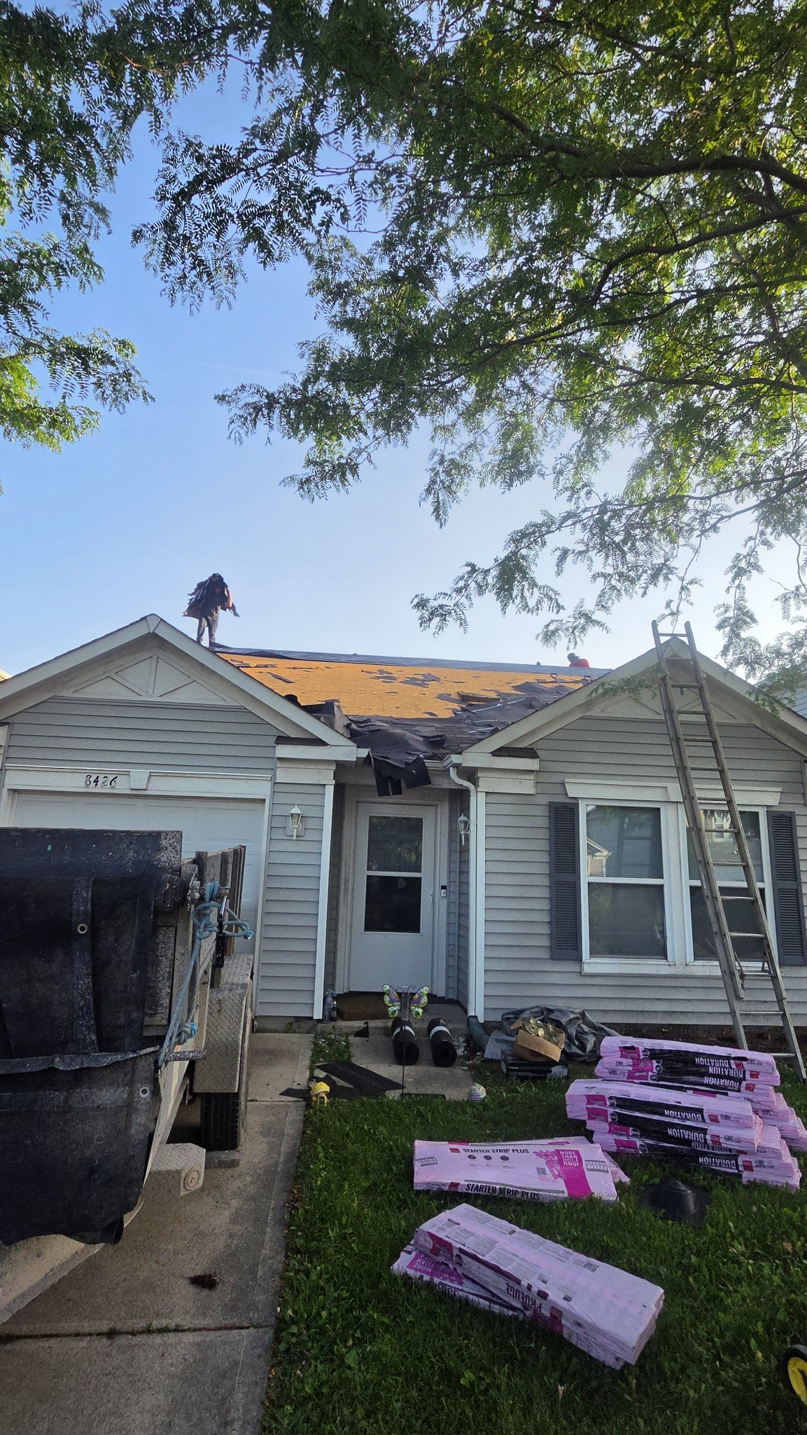 House at the start of a roof replacement with tear-off underway and shingle bundles on the lawn